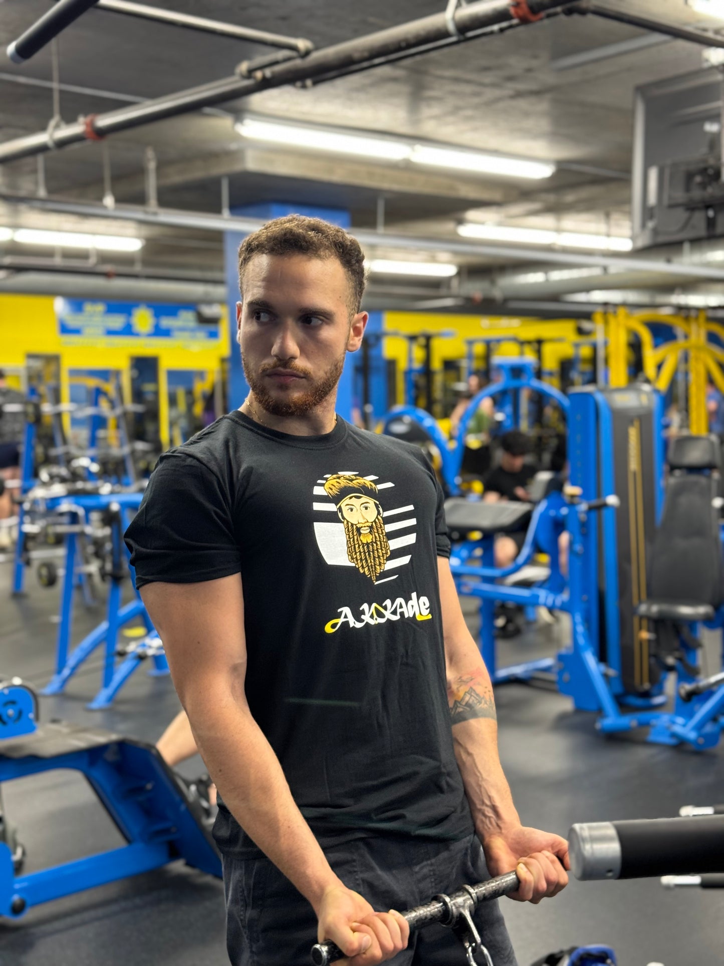 Man in a gym wearing a black t-shirt with a logo, holding a barbell.